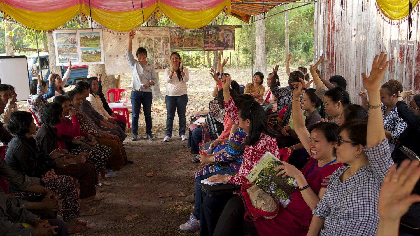People gather under a tent outdoors for a training workshop in Cambodia. Some participants have their hands raised as a response to moderators. Colorful banners and images hang from sides of the tent.